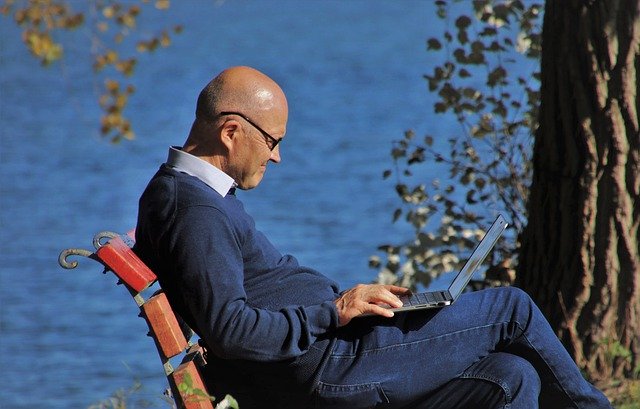 Muž bez vlasov sedí na lavičke v parku a pozerá do notebooku.jpg