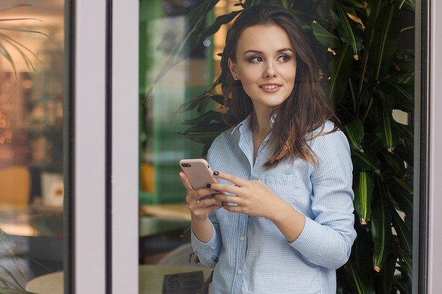 Žena stojí za sklenenou stenou, usmieva sa a píše na mobile